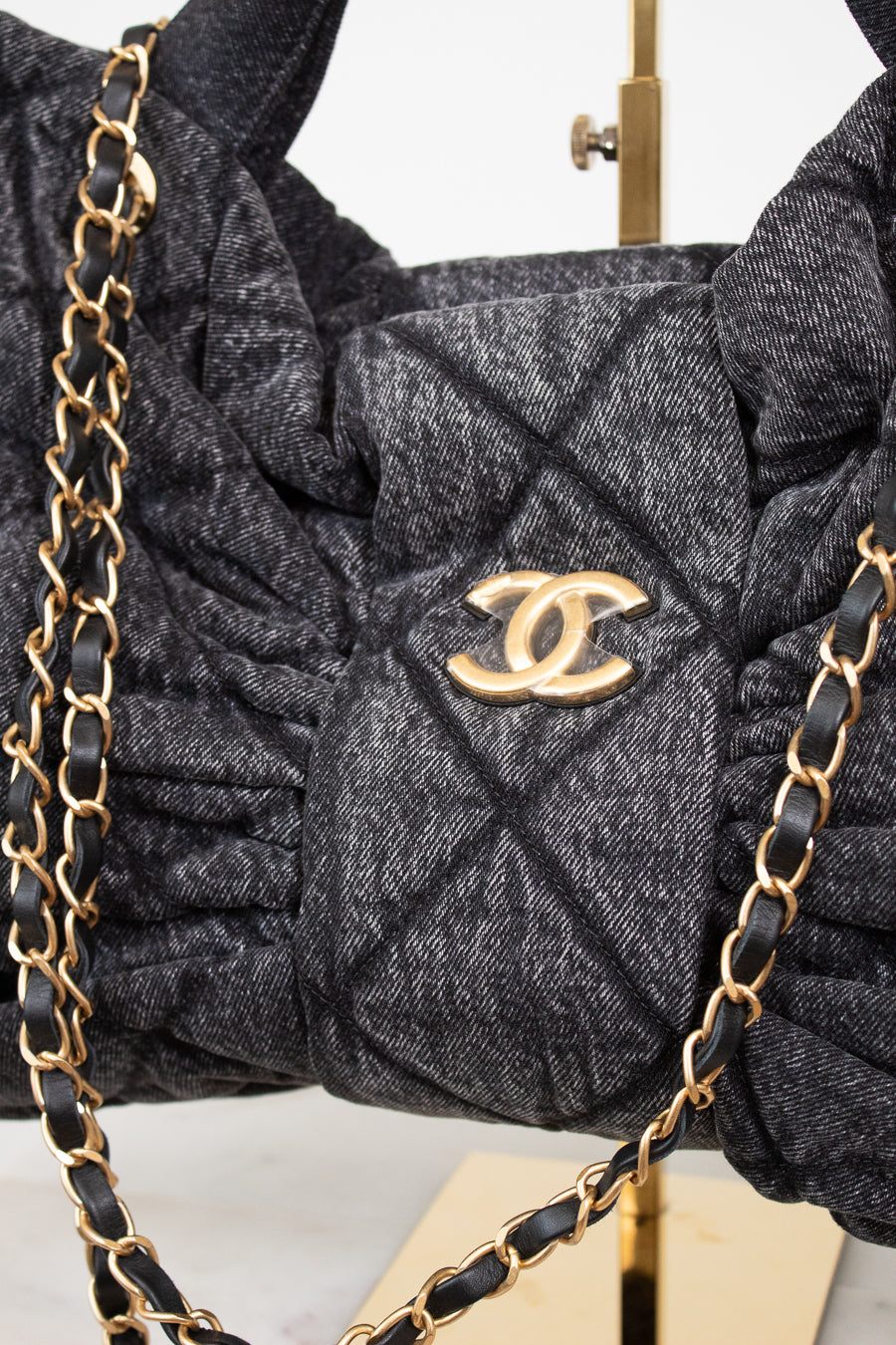 A close-up of a dark gray quilted Chanel shoulder bag with a gold-tone interlocking CC logo, featuring gathered detailing and a black-and-gold chain strap.
