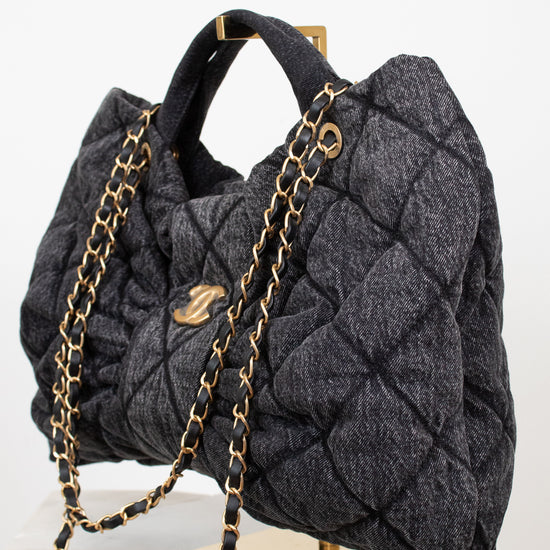 A dark gray quilted wool handbag with gold-tone chain straps and hardware, displayed on a gold stand over a white marble surface against a white background.