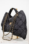 A dark gray quilted wool handbag with gold-tone chain straps and hardware, displayed on a gold stand over a white marble surface against a white background.