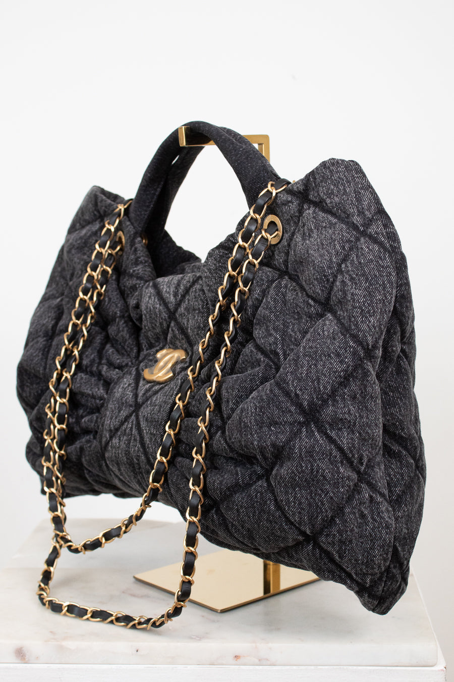A dark gray quilted wool handbag with gold-tone chain straps and hardware, displayed on a gold stand over a white marble surface against a white background.