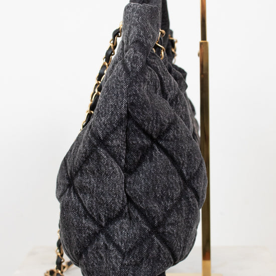 A dark gray quilted denim-like shoulder bag with gold-tone chain straps, displayed on a gold metal stand against a white background.