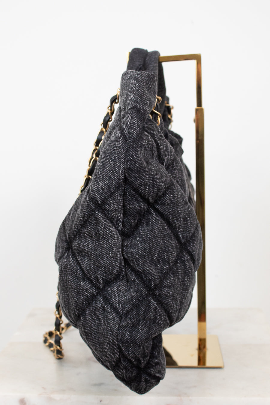 A dark gray quilted denim-like shoulder bag with gold-tone chain straps, displayed on a gold metal stand against a white background.