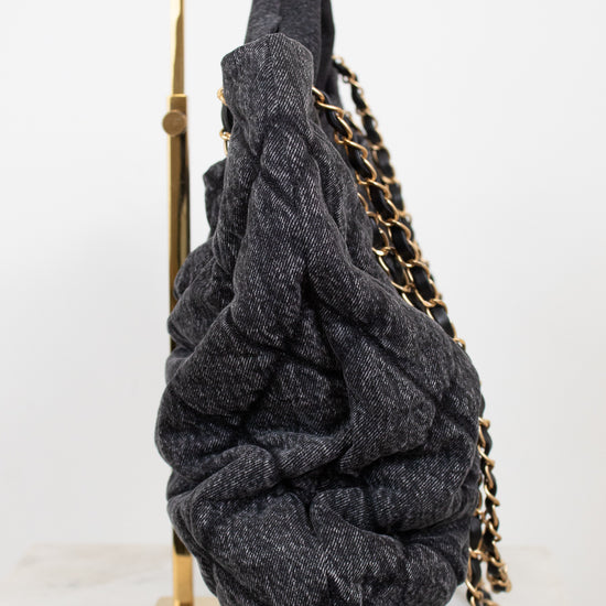 A dark gray denim-like, heavily textured shoulder bag with a gold chain strap, draped over a gold metal display stand against a white background.
