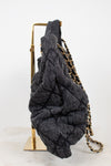 A dark gray denim-like, heavily textured shoulder bag with a gold chain strap, draped over a gold metal display stand against a white background.