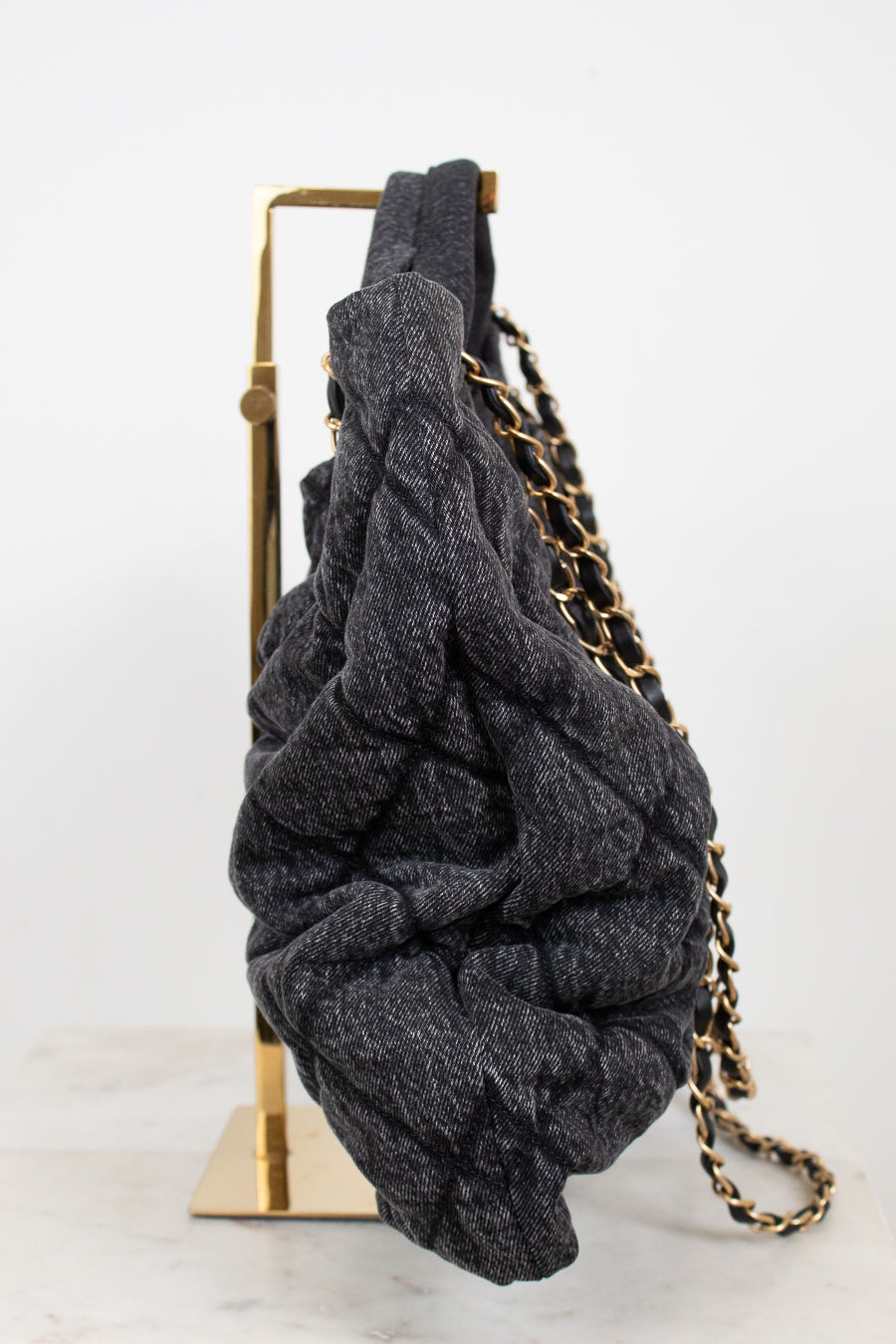 A dark gray denim-like, heavily textured shoulder bag with a gold chain strap, draped over a gold metal display stand against a white background.