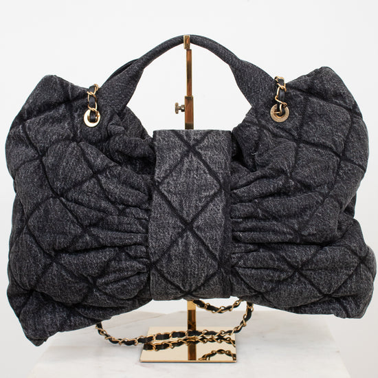 A dark gray quilted shoulder bag with gold-tone hardware, displayed on a gold stand atop a white marble block against a white background.