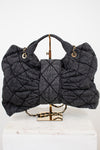 A dark gray quilted shoulder bag with gold-tone hardware, displayed on a gold stand atop a white marble block against a white background.