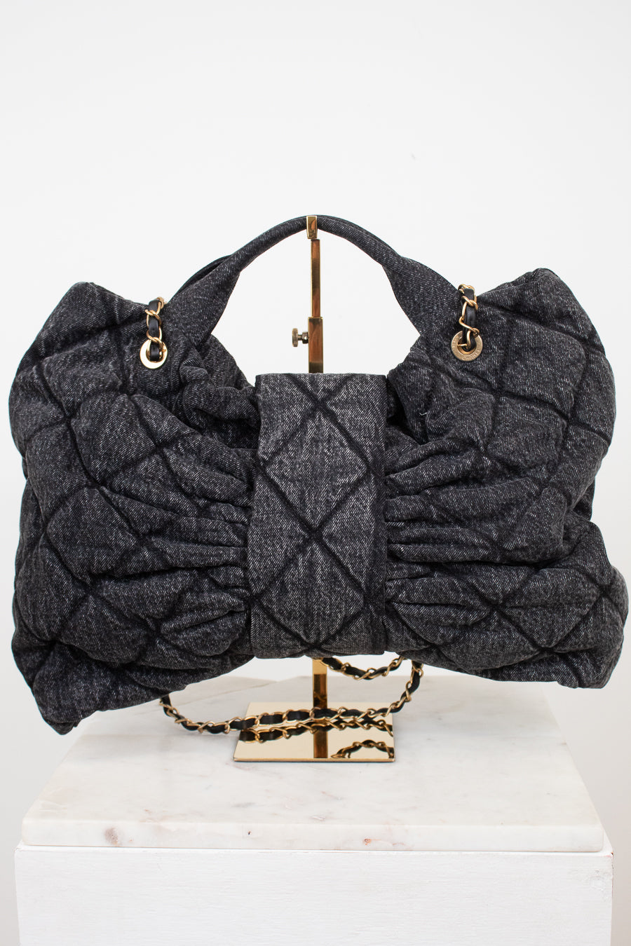A dark gray quilted shoulder bag with gold-tone hardware, displayed on a gold stand atop a white marble block against a white background.