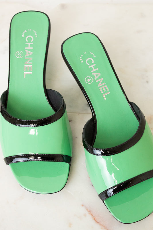 A pair of Chanel slide sandals in glossy mint green patent leather with black trim, displayed on a light marble surface. The insoles feature the white CHANEL logo and 'MADE IN ITALY' text.