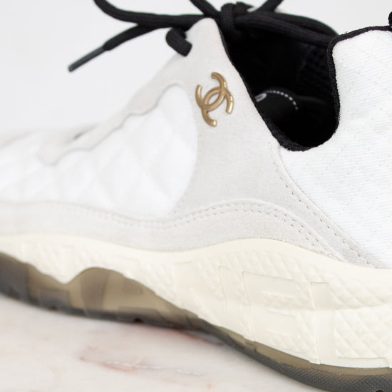 White sneaker with black laces and a gold logo on a light background