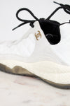 White sneaker with black laces and a gold logo on a light background