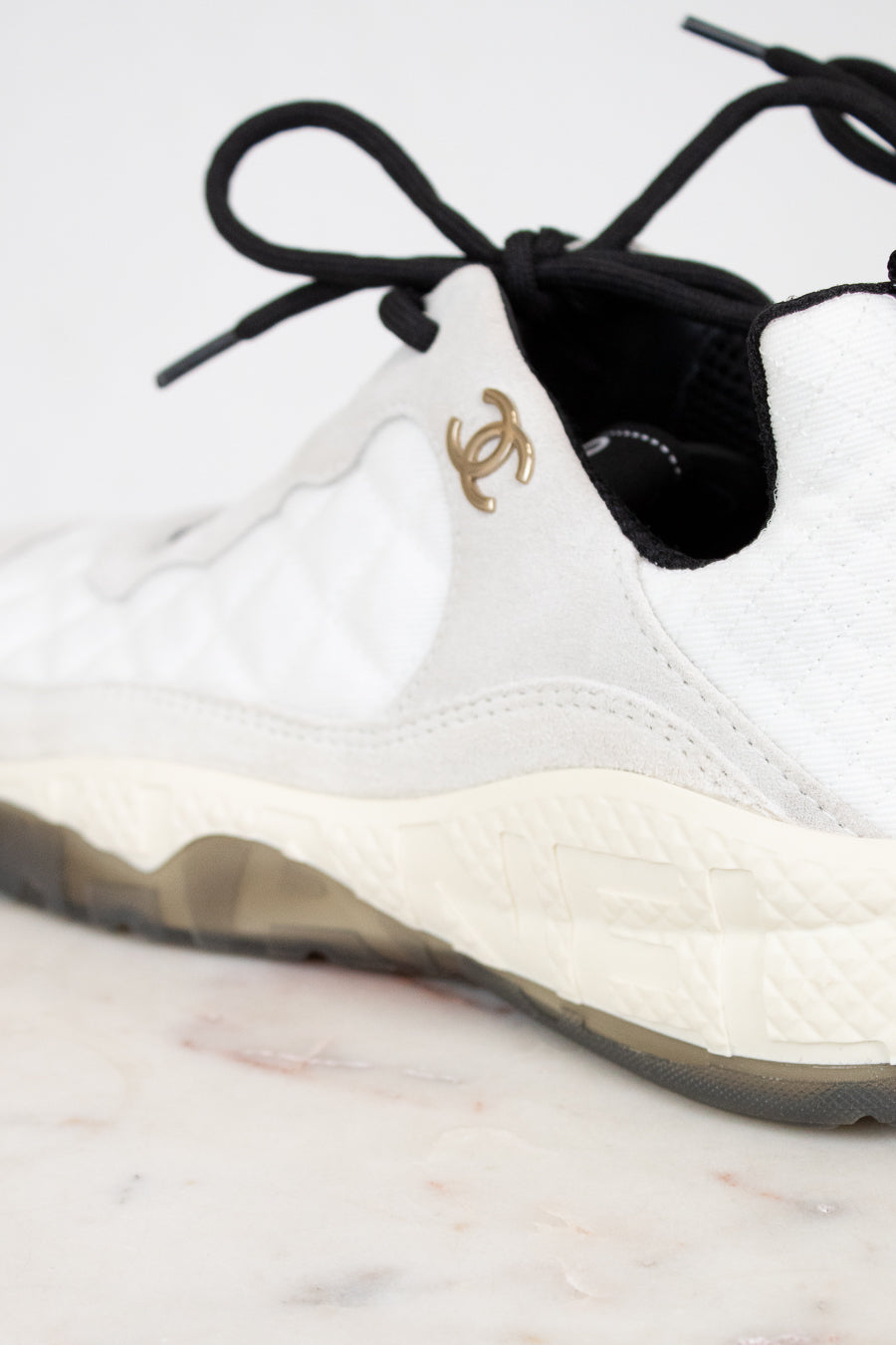 White sneaker with black laces and a gold logo on a light background