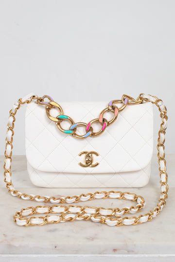 White quilted handbag with gold chain with multicolor details on strap help against a light gray background