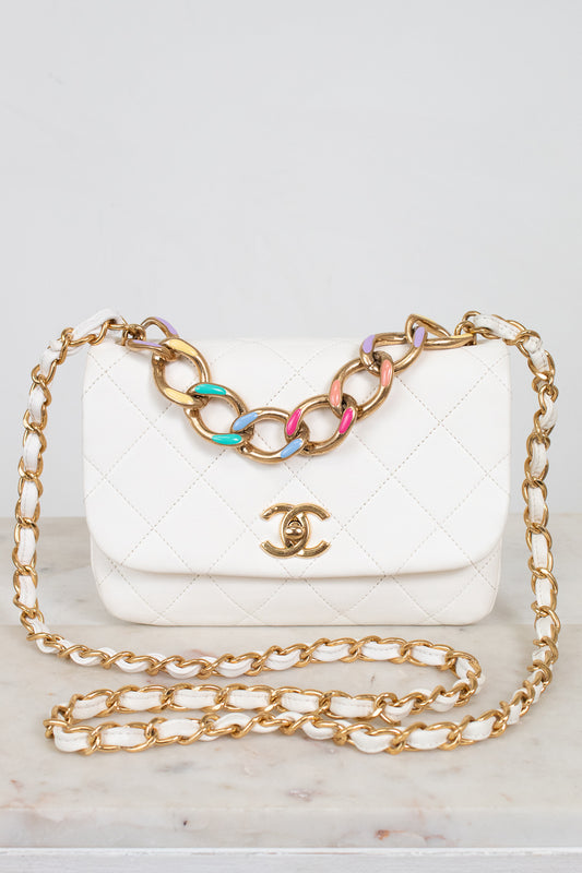 White quilted handbag with gold chain with multicolor details on strap help against a light gray background