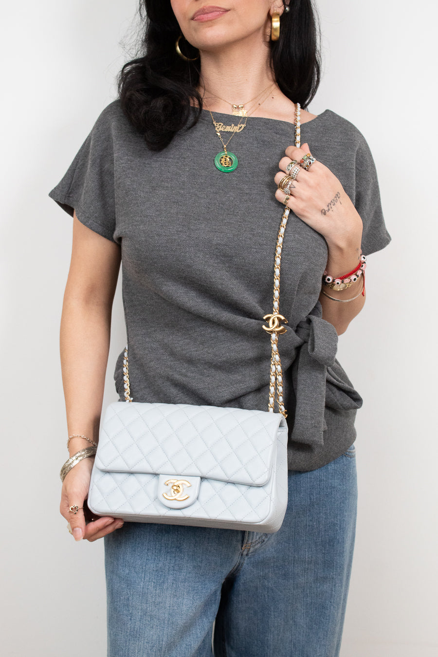 A woman wearing a gray t-shirt and blue jeans carries a light blue quilted Chanel shoulder bag with a gold chain strap and CC turn-lock closure.