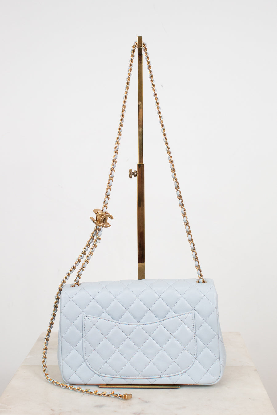 A light blue quilted leather shoulder bag with gold-tone chain straps and a gold interlocking CC logo charm, displayed on a stand against a white background.