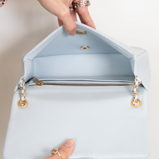 A light blue leather handbag shown open, with a person's hands holding the flap and bottom edge. The bag has gold-tone hardware, a zippered interior, and a chain strap against a plain off-white background.