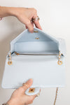 A light blue leather handbag shown open, with a person's hands holding the flap and bottom edge. The bag has gold-tone hardware, a zippered interior, and a chain strap against a plain off-white background.