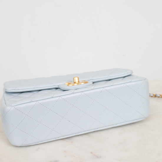 A light blue quilted leather handbag with a gold turn-lock clasp, resting on a white marble surface against a white background.