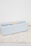 A light blue quilted leather handbag with a gold turn-lock clasp, resting on a white marble surface against a white background.