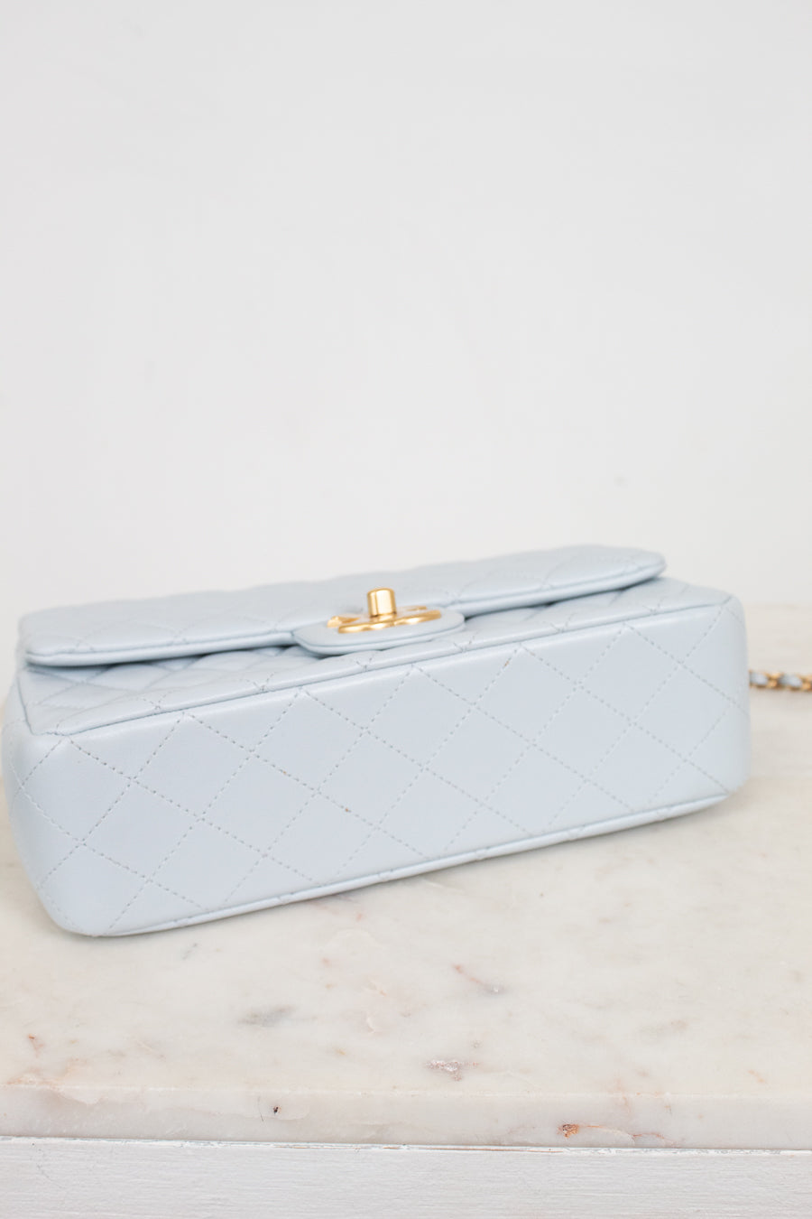 A light blue quilted leather handbag with a gold turn-lock clasp, resting on a white marble surface against a white background.