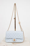 A light blue quilted leather shoulder bag with gold-tone hardware, displayed on a stand against a plain white background.
