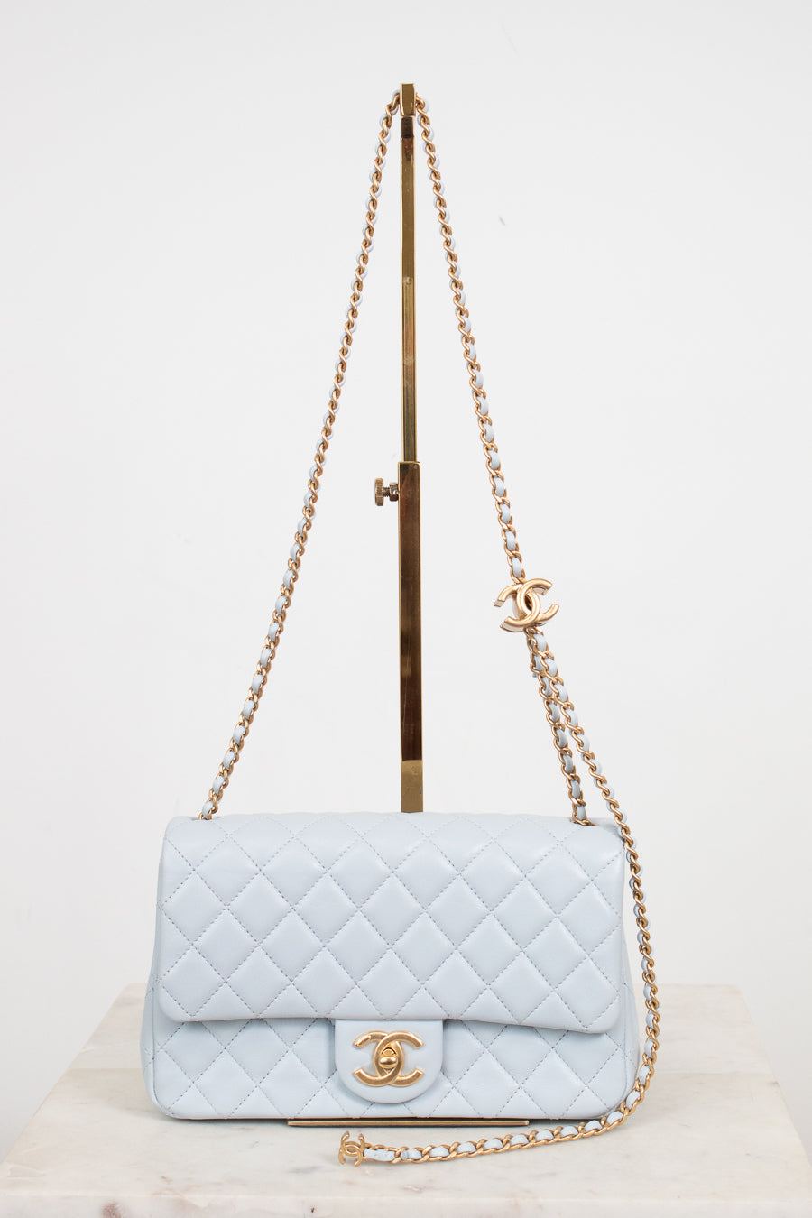 A light blue quilted leather shoulder bag with gold-tone hardware, displayed on a stand against a plain white background.