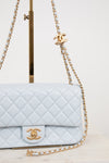 A light blue quilted Chanel flap bag with gold-tone hardware, shown on a display stand against a clean white background.