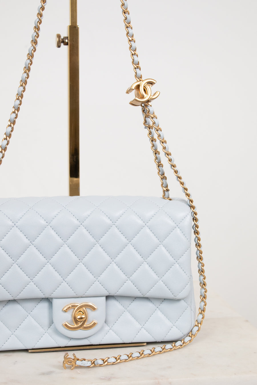 A light blue quilted Chanel flap bag with gold-tone hardware, shown on a display stand against a clean white background.