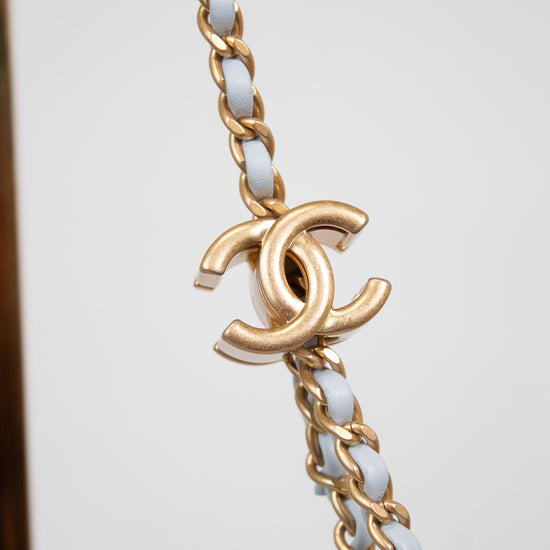 A close-up of a gold-tone chain bracelet featuring interwoven white faux-leather links and a prominent interlocking CC logo charm, characteristic of Chanel.