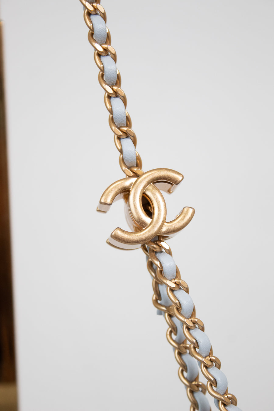 A close-up of a gold-tone chain bracelet featuring interwoven white faux-leather links and a prominent interlocking CC logo charm, characteristic of Chanel.