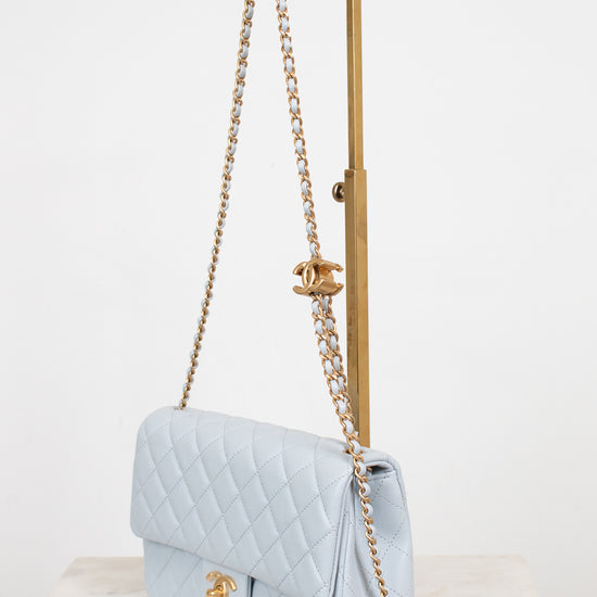 A light blue quilted Chanel shoulder bag with gold-tone hardware, displayed on a gold metal stand over a marble surface against a white background.