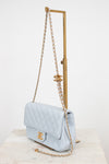 A light blue quilted Chanel shoulder bag with gold-tone hardware, displayed on a gold metal stand over a marble surface against a white background.