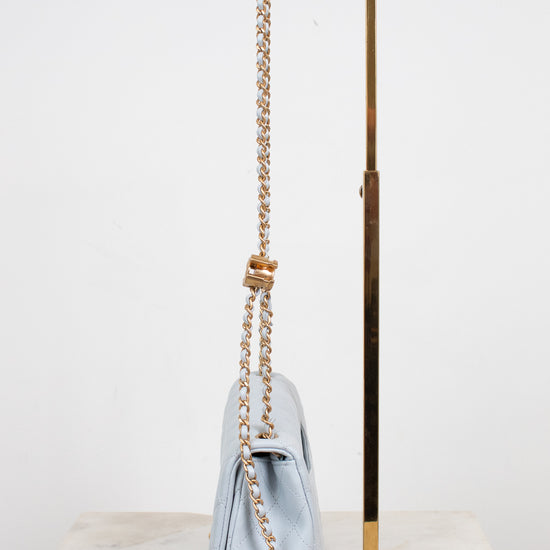 A light blue quilted leather shoulder bag with a gold chain strap and a gold knotted detail, displayed on a marble surface next to a gold metal stand.