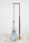 A light blue quilted leather shoulder bag with a gold chain strap and a gold knotted detail, displayed on a marble surface next to a gold metal stand.