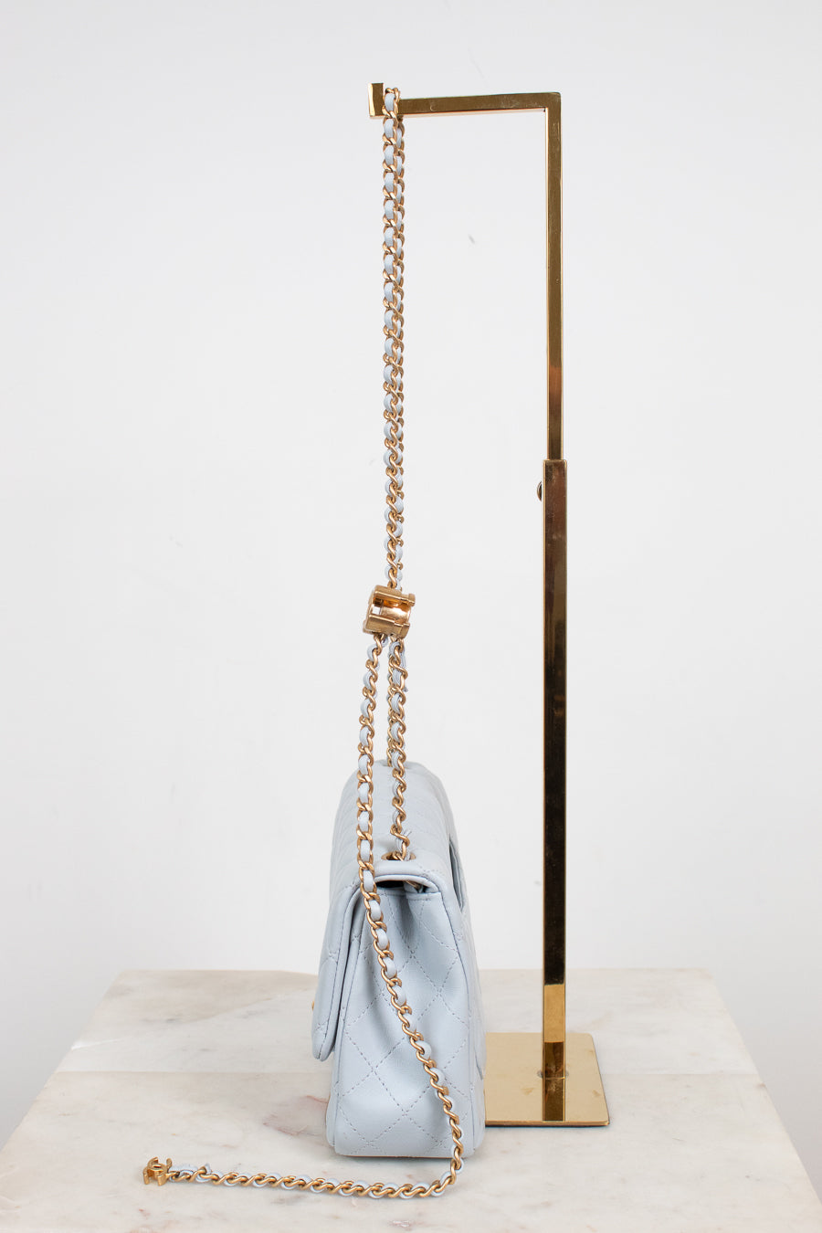 A light blue quilted leather shoulder bag with a gold chain strap and a gold knotted detail, displayed on a marble surface next to a gold metal stand.