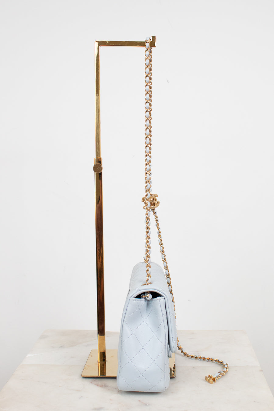 A light blue quilted leather shoulder bag with a gold chain strap, displayed on a gold metal stand against a white background.
