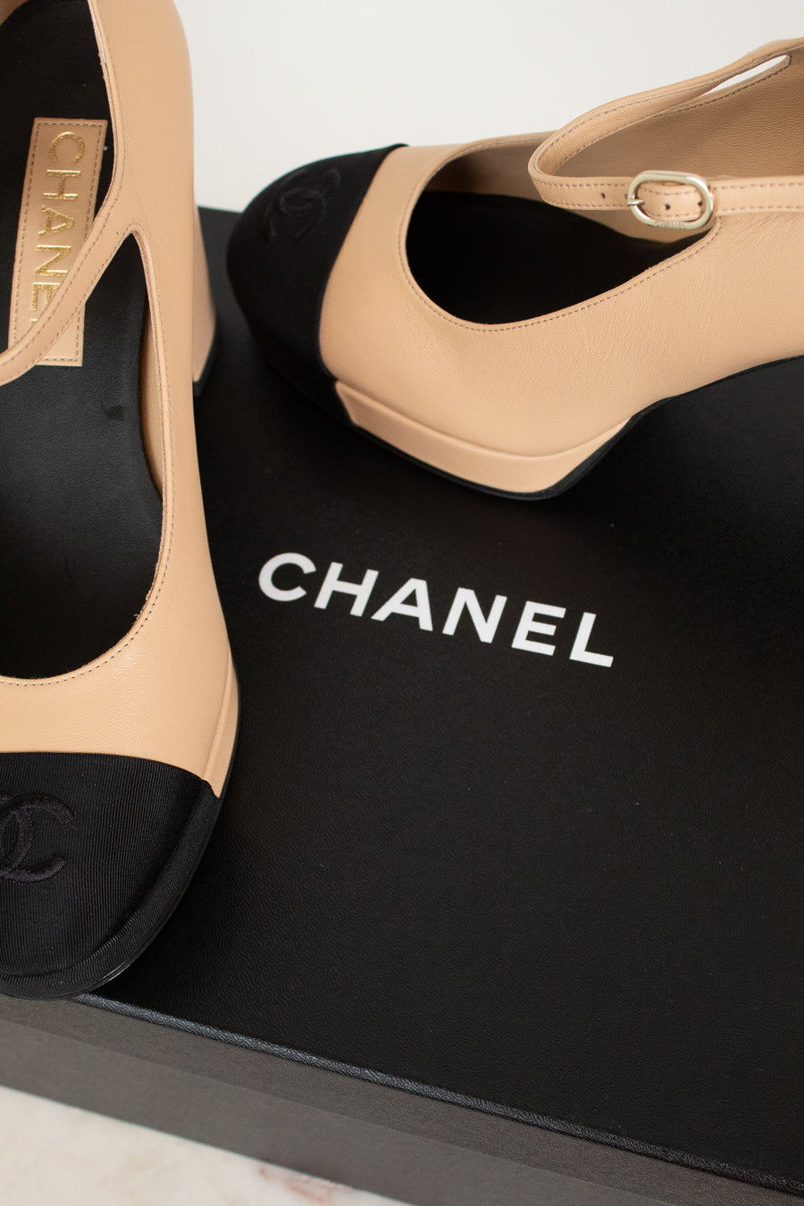 Beige and black Chanel high-heeled shoes on a black box with the Chanel logo.