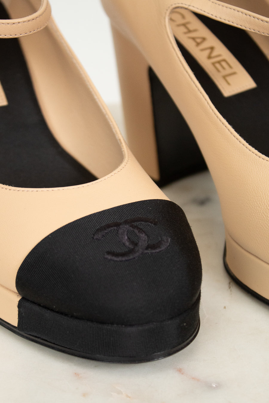 Close-up of beige high-heeled shoes with black featuring Chanel logo on a light background.