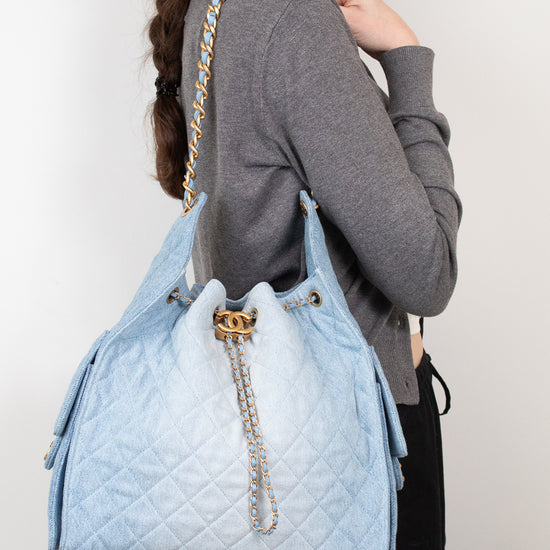 A woman wearing a gray cardigan and black pants carries a large light blue quilted shoulder bag with gold-tone chain detailing and a gold double-C logo clasp, shown from the side against a plain light background.