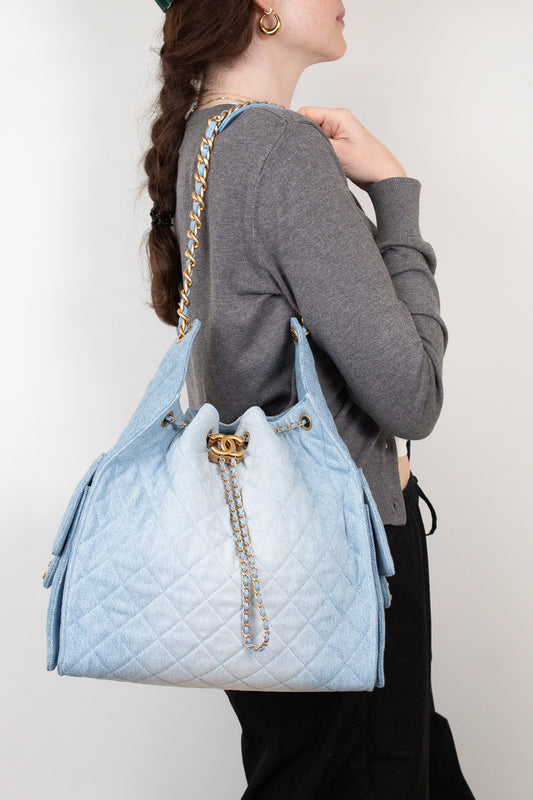A woman wearing a gray cardigan and black pants carries a large light blue quilted shoulder bag with gold-tone chain detailing and a gold double-C logo clasp, shown from the side against a plain light background.