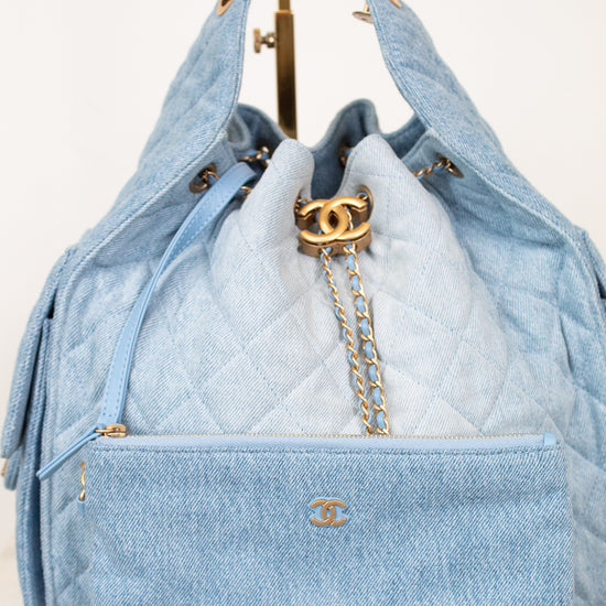 A light blue quilted Chanel handbag with gold-tone hardware, shown with a matching zip pouch in front and chain straps hanging above on a display stand.
