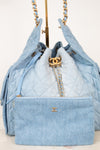 A light blue quilted Chanel handbag with gold-tone hardware, shown with a matching zip pouch in front and chain straps hanging above on a display stand.