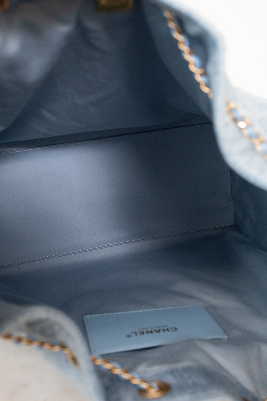 Close-up of the interior of a Chanel handbag in a muted blue-gray color, showing the branded CHANEL interior card and a gold-tone chain strap.