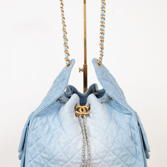 A light blue quilted drawstring handbag with gold-tone chain straps and a gold interlocking CC clasp, displayed on a stand against a white background.