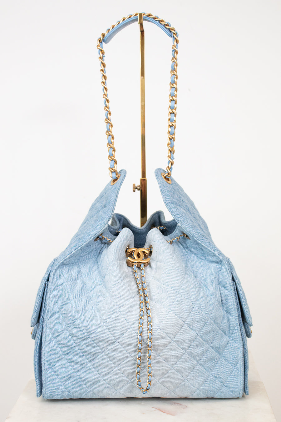 A light blue quilted drawstring handbag with gold-tone chain straps and a gold interlocking CC clasp, displayed on a stand against a white background.