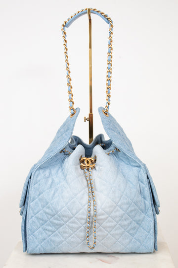 A light blue quilted drawstring handbag with gold-tone chain straps and a gold interlocking CC clasp, displayed on a stand against a white background.