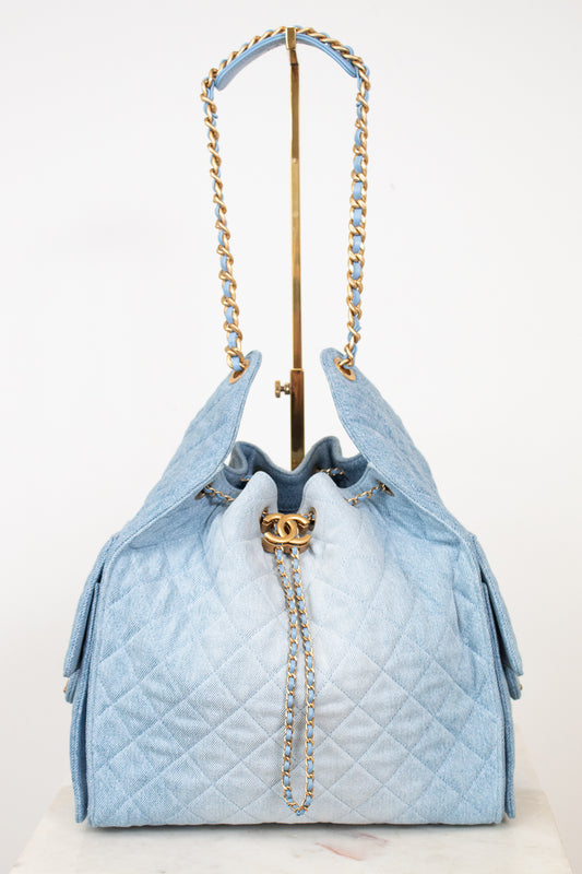 A light blue quilted drawstring handbag with gold-tone chain straps and a gold interlocking CC clasp, displayed on a stand against a white background.