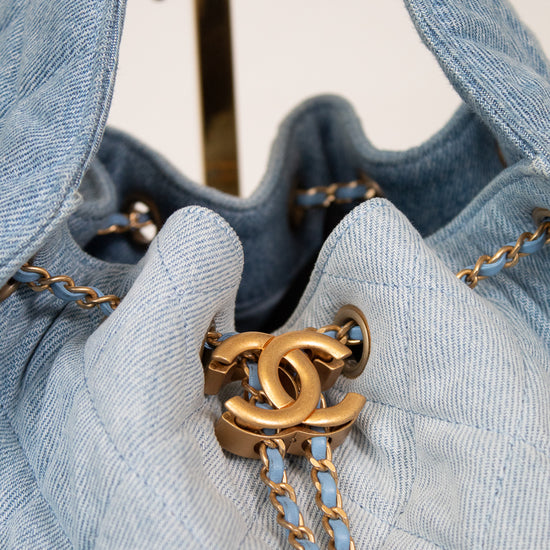 A close-up of a light blue denim drawstring bag with gold-tone hardware, featuring a prominent interlocking CC logo and a chain strap on a neutral background.
