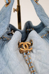 A close-up of a light blue denim drawstring bag with gold-tone hardware, featuring a prominent interlocking CC logo and a chain strap on a neutral background.